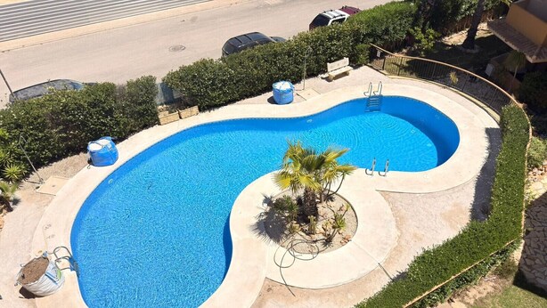 A swimming pool with a curved design, surrounded by a paved deck and greenery. - Recoleta Penthouse Punta Prima (Torrevieja)