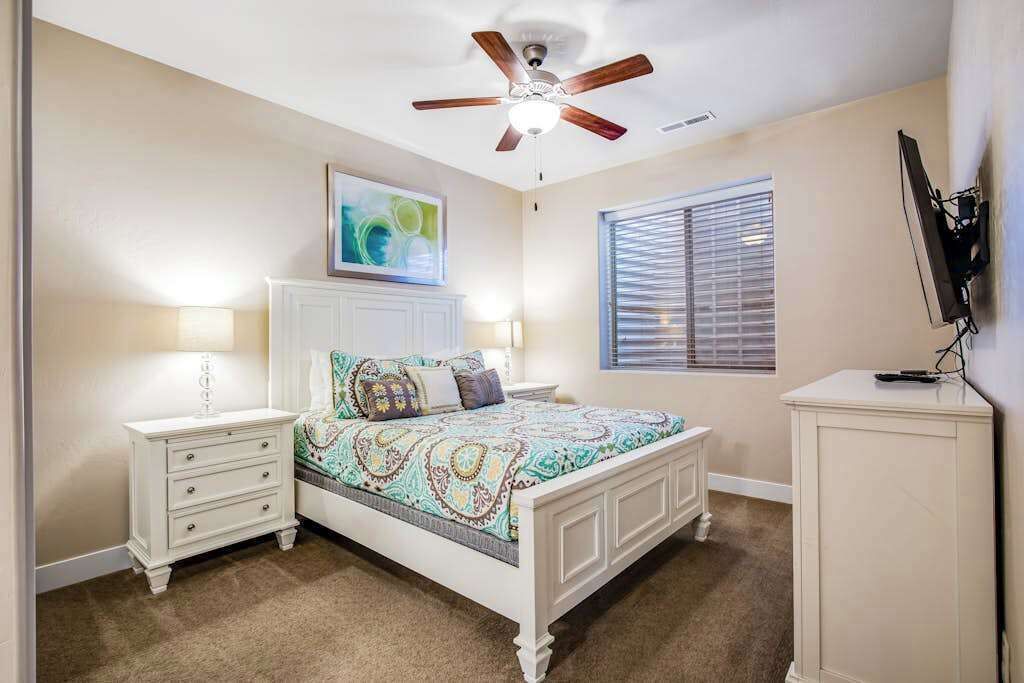 A bedroom with a bed, two nightstands, a ceiling fan, a window with blinds, and a TV on a stand.