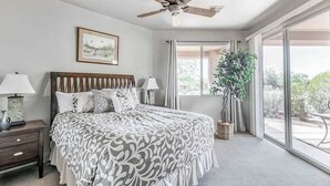 Quarto com cama, mesas de cabeceira, ventoinha no teto, janela com cortinas e um vaso de plantas.