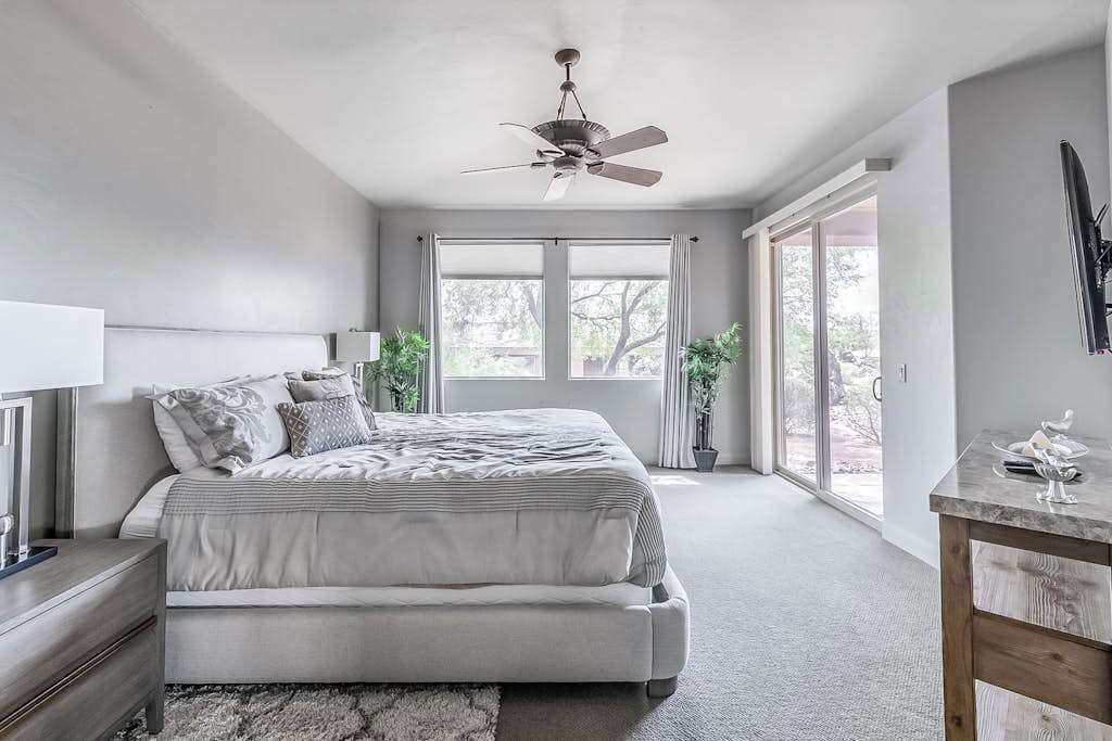 Un dormitorio con cama, mesita de noche, ventilador de techo, ventanas amplias y vistas al exterior.