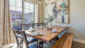 Un comedor con una mesa de madera dispuesta para seis, sillas, un banco, una araña y un cuadro abstracto grande en la pared.