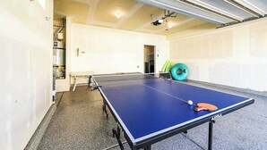 Una mesa de ping pong con una pelota y una paleta, una pelota de ejercicios verde, y una puerta blanca en una habitación con suelo de baldosas.
