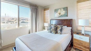 Ein Hotelzimmer mit einem großen Bett, einem Nachttisch mit Lampe, einem Fenster mit Vorhängen und einem an der Wand befestigten Kunstwerk.