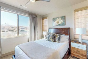 Ein Hotelzimmer mit einem großen Bett, einem Nachttisch mit Lampe, einem Fenster mit Vorhängen und einem an der Wand befestigten Kunstwerk.