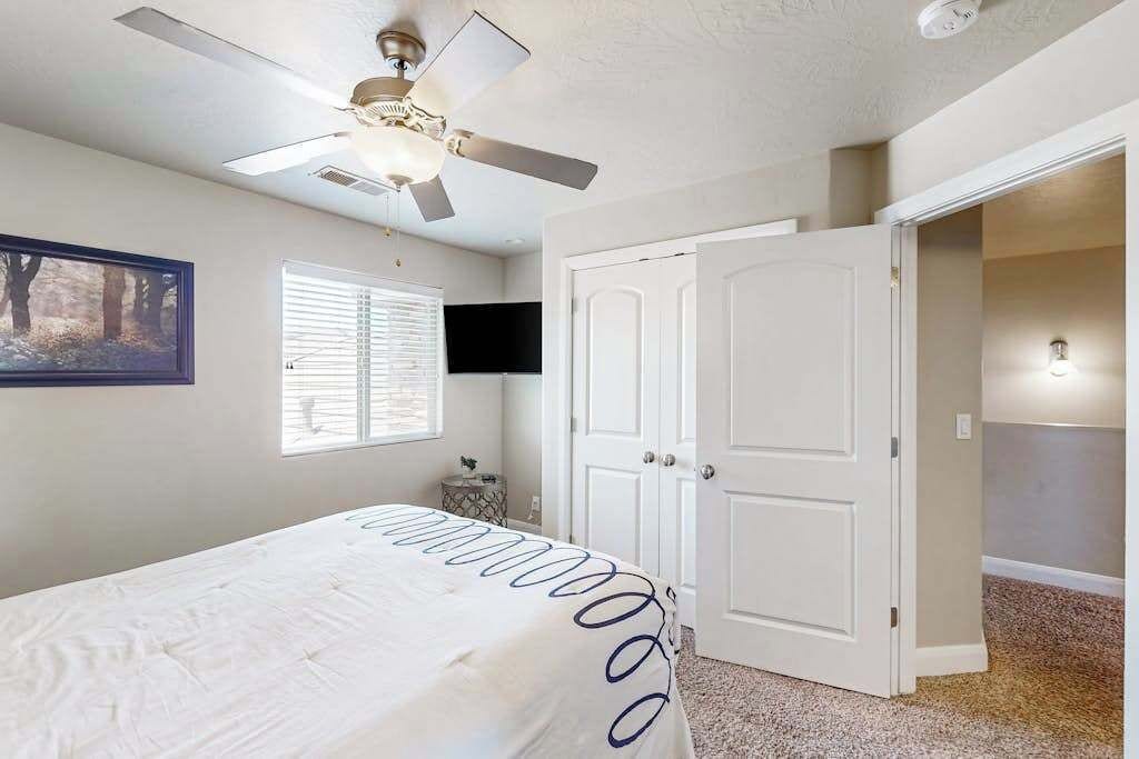 A bedroom with a bed, a ceiling fan, a TV, a door, and a window with blinds.