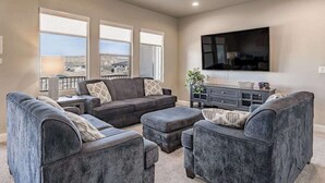 A living room with a large flat-screen TV, grey upholstered furniture, and a view through large windows. - 6BR Spacious townhouse with a pool & lazy river (Hurricane)