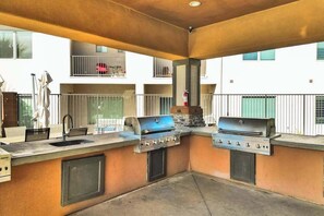 Ein Außenbereich mit Küche, ausgestattet mit zwei Gasgrills, einem Spülbecken und einem Fenster mit Blick auf ein Wohngebäude.