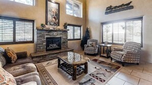 A spacious living room with a stone fireplace, large windows, and a high ceiling with exposed beams. - Spacious 5BR Mountainview Zion National Park (Leeds)