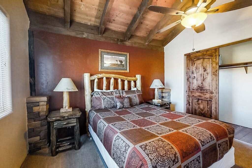 Ein Schlafzimmer mit einem hölzernen Kopfende, einer gemusterten Bettdecke, zwei Nachttischlampen, einer rustikalen Tür und einem Deckenventilator.