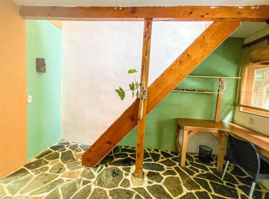 Una habitación con techo inclinado, estructura de vigas de madera, suelo de piedra, una mesa pequeña y una silla.