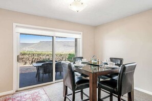 Uma sala de jantar com uma mesa posta para quatro pessoas, uma porta de vidro deslizante que dá para um pátio exterior, e vista para uma paisagem montanhosa.