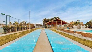 Zona de piscina con un camino que lleva a un edificio, rodeada por una valla y vegetación.