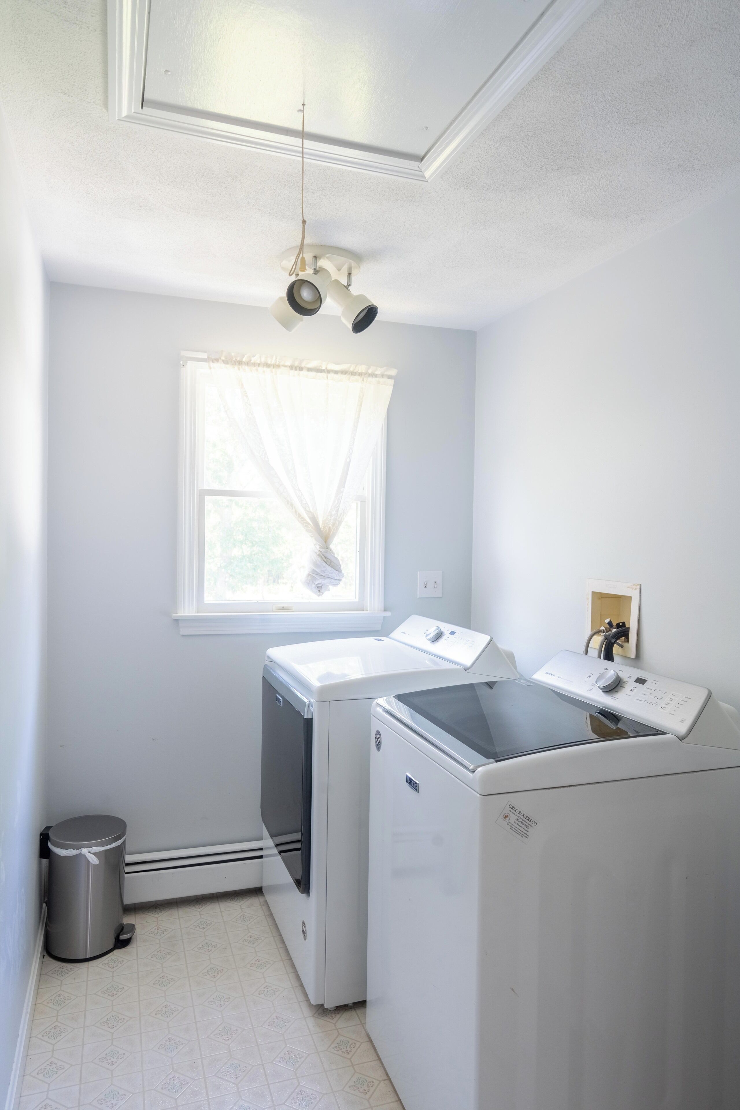A laundry room with two washing machines, a window with sheer curtains, and a ceiling light fixture.