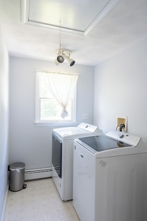 A laundry room with two washing machines, a window with sheer curtains, and a ceiling light fixture.