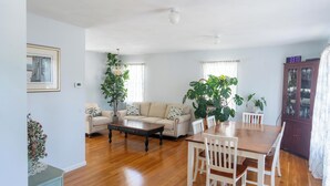A spacious living room with a wooden floor, a beige sofa, a coffee table, and a dining table with chairs. There are potted plants and a framed picture on the walls.