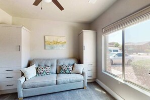 A room with a grey sofa, a ceiling fan, a window with a view of the outside, and a painting on the wall. - Townhouse with resort amenities, private hot tub (Hurricane)