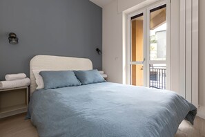 A bedroom with a bed, pillows, a nightstand, and a window with a view. - Blue Two-Room Apartment with a Terrace, 5 Minutes from the Station (Bergamo)