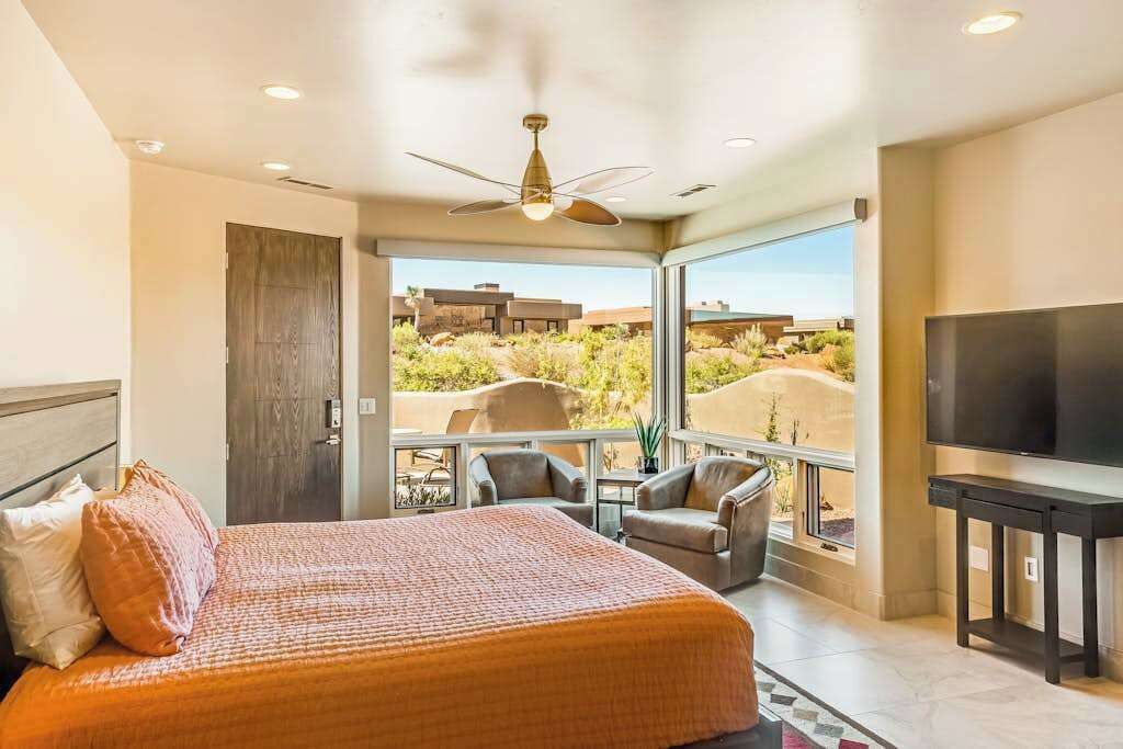 A bedroom with a bed, a TV, a ceiling fan, and a balcony with a view of the desert.
