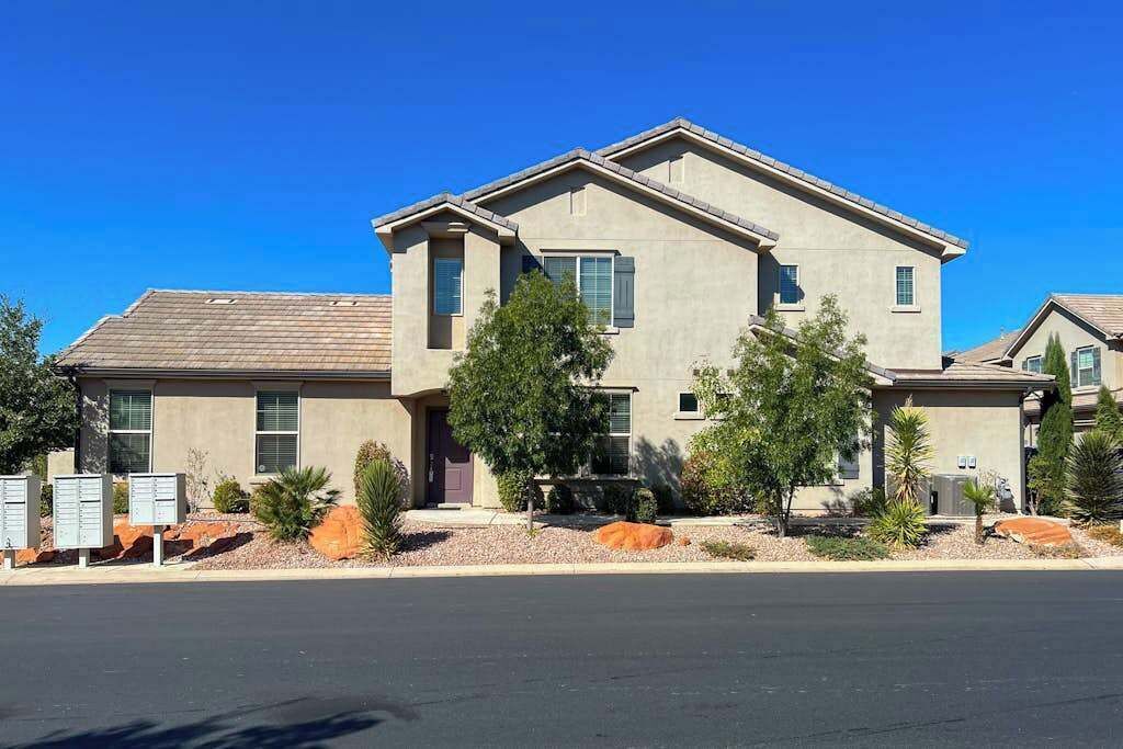 A two-story residential building with a flat roof, multiple windows, and a landscaped front yard with shrubs and a small tree.