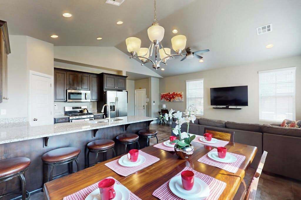 A modern kitchen with a central island, bar stools, a dining table set for four, and a living area with a sofa and TV.