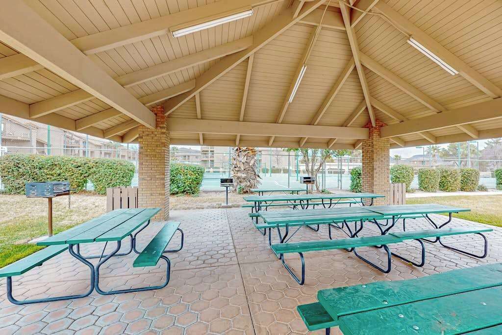 An outdoor covered picnic area with multiple green picnic tables and a brick pillar.