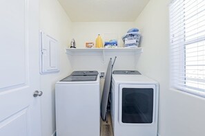 Uma lavanderia com duas máquinas de lavar e secadoras brancas, uma prateleira com produtos de lavagem e uma porta com interruptor de luz.
