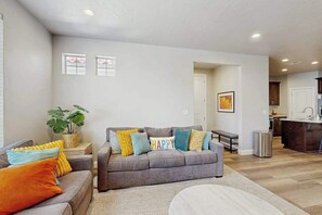 Ein modernes Wohnzimmer mit einem grauen Sofa, farbenfrohen Kissen, einer Bank und einem Essbereich im Hintergrund.