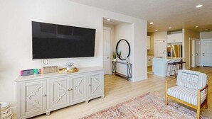 Una sala de estar con televisión de pantalla plana, un aparador blanco, un tapete estampado y una silla de madera.