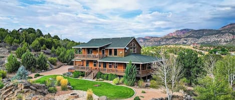 A two-story wooden house with a large deck, surrounded by a landscaped garden and rocky terrain.