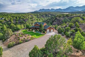A large house with a well-maintained lawn and driveway, surrounded by dense vegetation and mountains in the background. - 4BR home - wrap around porch, fire pit & hot tub (New Harmony)