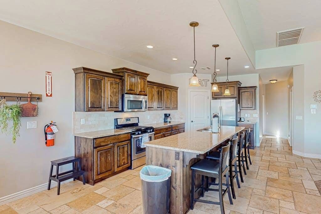 A kitchen with wooden cabinets, a central island with bar stools, a stainless steel refrigerator, and a hanging light fixture.