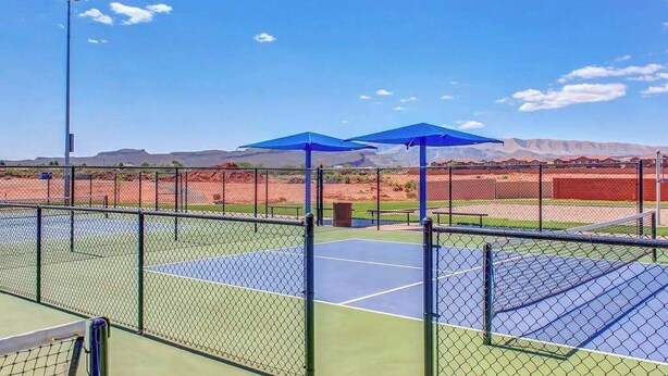 Tenniskenttä, jossa on metallinen verkkoaita, siniset aurinkovarjot ja kirkas taivas.