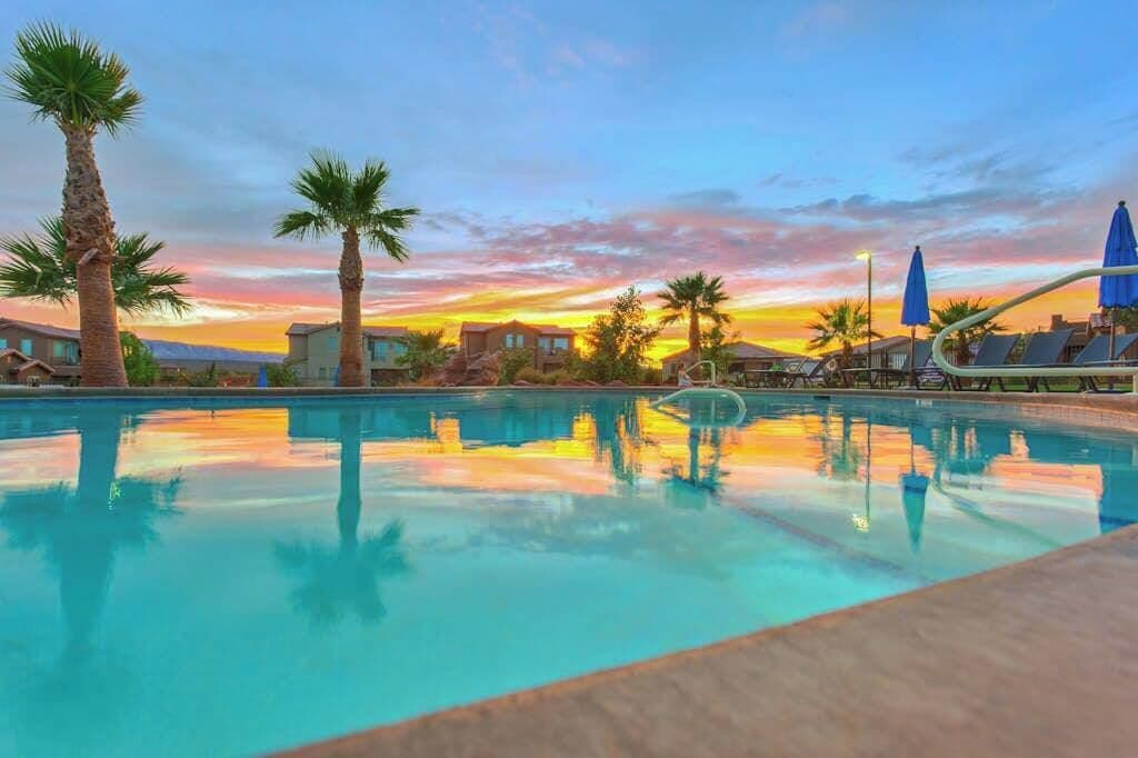 A swimming pool with palm trees and lounge chairs, reflecting a sunset sky.