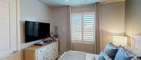 Une chambre à coucher comprenant un lit, un téléviseur, un ventilateur de plafond et une fenêtre avec des stores.