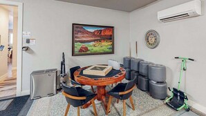 Um quarto com uma mesa de madeira redonda, cadeiras pretas, um quadro de uma paisagem, um aparelho de ar condicionado, um relógio e bolas de exercício.