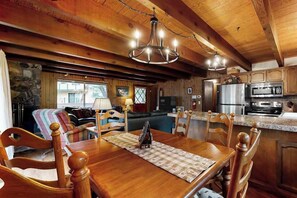 A wooden interior with a dining table, chairs, a chandelier, and a kitchen area with appliances.