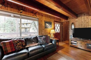 A cozy living room with a leather sofa, wooden walls, a television, and a window with a view of trees.