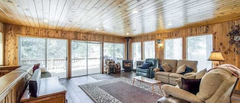 A spacious room with wooden walls and ceiling, a large window, a fireplace, and a seating area with a sofa and armchairs.