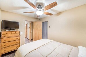 Une chambre à coucher avec un lit, une commode en bois, un téléviseur posé dessus et un ventilateur de plafond.