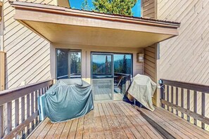 Eine Holzterrasse mit überdachter Sitzgruppe, eine Glastür, die auf einen Balkon führt, und Blick auf Bäume und Berge.