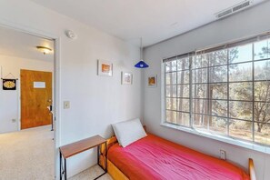 Ein Schlafzimmer mit einem Einzelbett, einem Fenster mit Blick auf Bäume, einem Nachttisch und einer Tür zu einem anderen Raum.