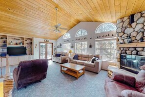 A spacious living room with a high ceiling, wooden beams, and a stone fireplace. There are multiple seating areas and a television. - Escape to Yosemite - Pine Mountain Lake (Groveland)