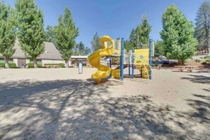 Ein Spielplatz mit einer gelben Rutsche und blauen Elementen, umgeben von Bäumen und einem Gebäude im Hintergrund.