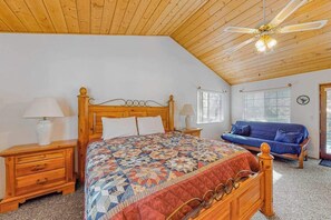 A bedroom with a wooden ceiling, a bed with a quilt, a nightstand, a lamp, a blue sofa, and a window. - Escape to Yosemite - Pine Mountain Lake (Groveland)