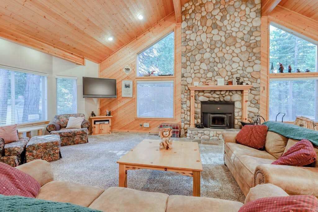 A cozy living room with a stone fireplace, wooden furniture, and large windows.