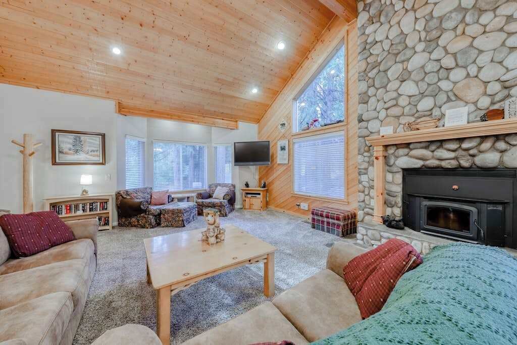 A cozy living room with a stone fireplace, wooden ceiling, and large windows.