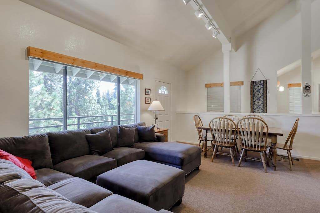 A living room with a sectional sofa, a dining table with chairs, and large windows.