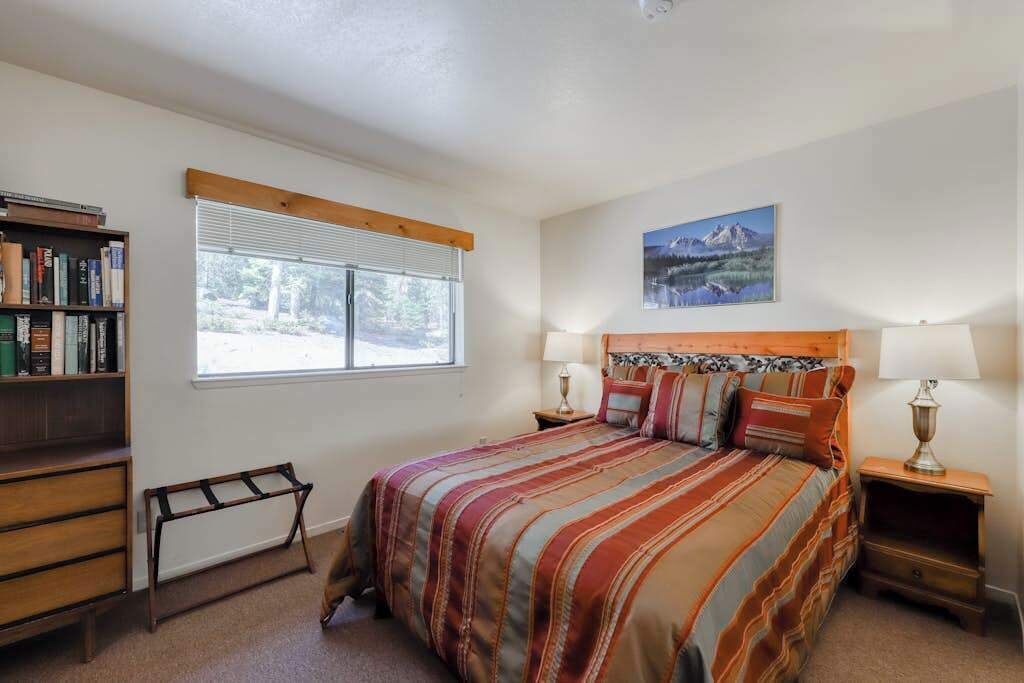 Un dormitorio con cama, mesitas de noche, una estantería, ventana con persianas y un cuadro en la pared.