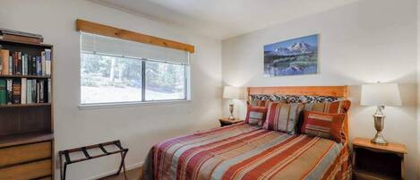 Un dormitorio con cama, mesitas de noche, una estantería, ventana con persianas y un cuadro en la pared.
