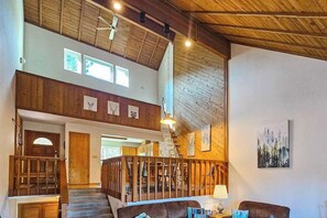 Een ruim interieur met een hoge plafond, houten balken, een trap en een zithoek met een lamp.